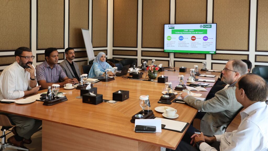 Meeting participants seated around a conference table during a presentation, with a screen displaying a timeline and partnership information.
